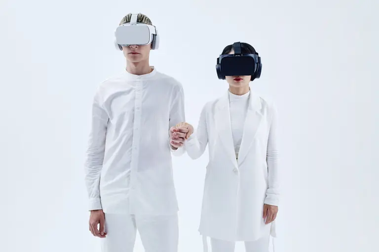 Two people in white attire wearing virtual reality headsets and holding hands, embracing technology.