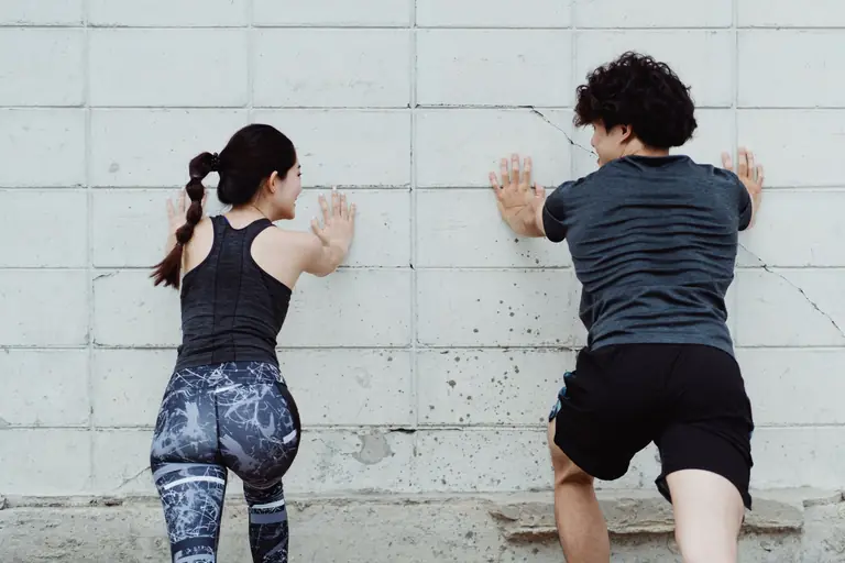 A man and woman push against a wall during an outdoor workout session, focusing on strength and fitness.