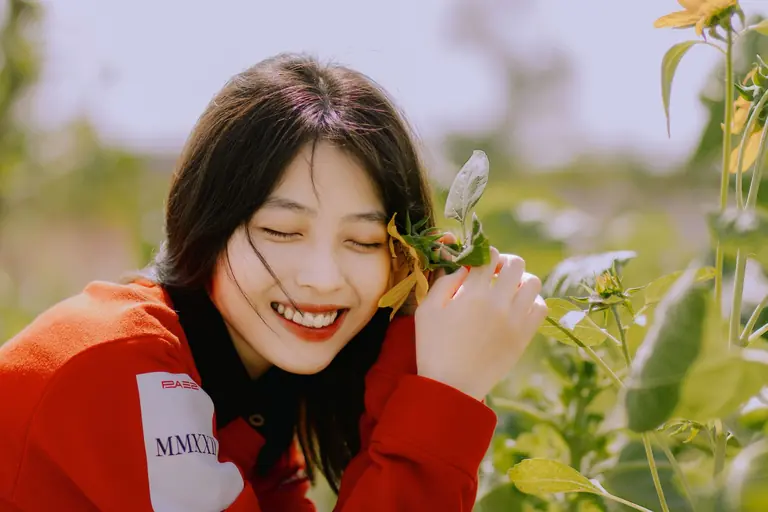 A smiling woman in a red shirt enjoys a sunny day among green plants and sunflowers.
