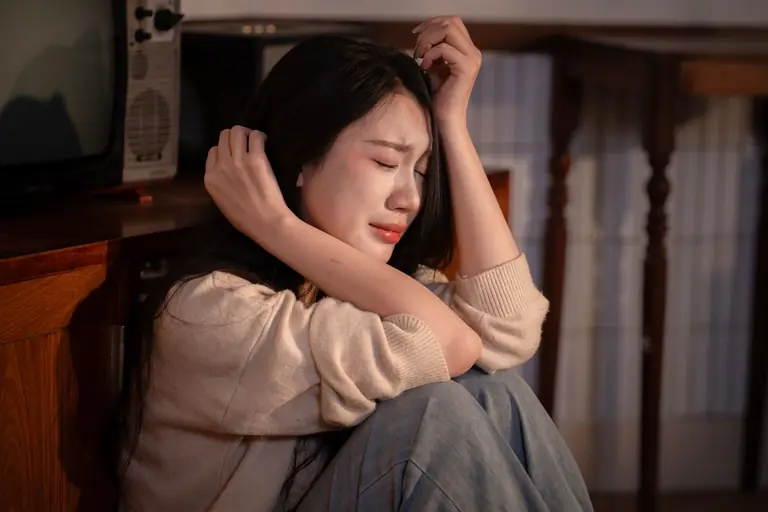 A young woman sitting indoors, expressing deep emotional distress.