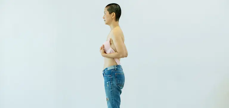 Side view of mature female putting pink bra to chest after breast removal surgery from cancer while standing in bright studio on white background