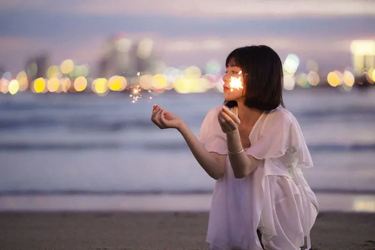 A young woman enjoys sparklers on the beach at twilight with city lights in the background.