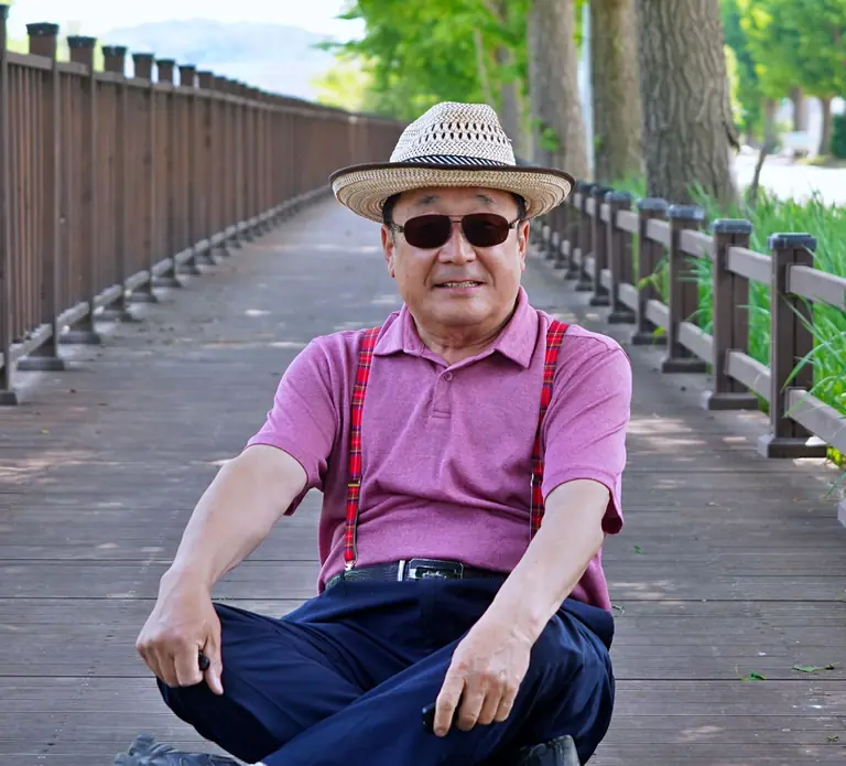 A senior man in a straw hat sits on a wooden path, enjoying a sunny day outdoors.