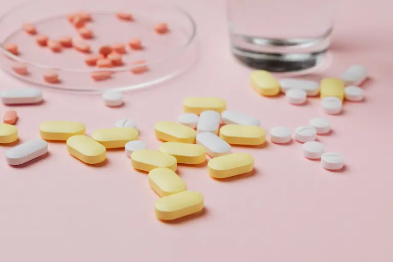 A close-up of various pills and tablets on a pink background with a glass of water.